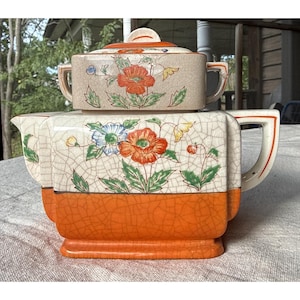 Pode incluir: Um bule de cerâmica quadrado com tampa, apresentando uma secção superior de cor creme com vidrado craquelado e um padrão floral de flores laranja, azuis e amarelas com folhas verdes, e uma secção inferior laranja sólida craquelada.