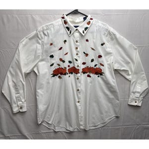 Puede incluir: Camisa blanca abotonada con mangas largas. La camisa presenta calabazas naranjas bordadas y hojas coloridas. El cuello también está decorado con hojas bordadas. La camisa tiene botones de madera y puños abotonados.