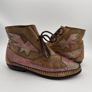 May include: A pair of brown leather ankle boots with pink and iridescent lightning bolt and scale pattern accents. The boots have brown laces and a stitched sole, offering a unique and stylish design.