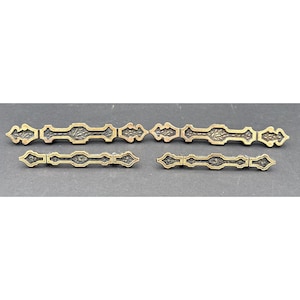 May include: A collection of antique brass drawer pulls with an ornate, symmetrical design. The pulls have a dark, aged finish and are arranged on a dark gray surface. The decorative hardware features a repeating pattern of stylized floral and geometric elements.