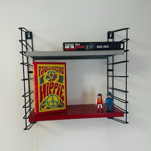 May include: A black metal shelf unit with two shelves. The top shelf is grey and holds a book titled "Sharon Tate and the Manson Murders" by Greg King. The bottom shelf is red and holds a book titled "Hippie" by Paulo Coelho and two small plastic figures.