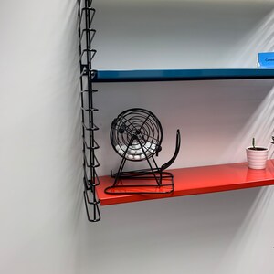 May include: A black metal lottery cage with white balls inside sits on a red shelf. The shelf is part of a black metal shelving unit.