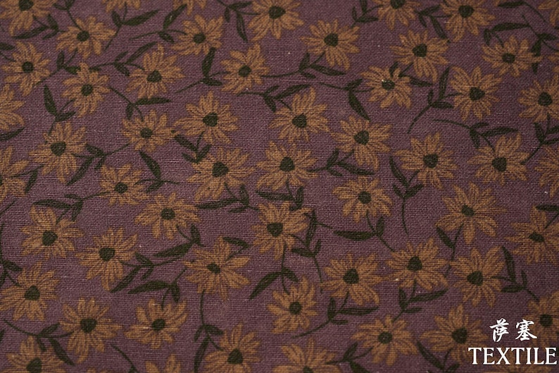 Peut inclure: Gros plan sur un tissu avec un motif floral r&eacute;p&eacute;t&eacute;. Les fleurs sont jaune dor&eacute; avec des centres fonc&eacute;s, sur un fond violet fonc&eacute;. Des feuilles et des tiges vert fonc&eacute; s'entrelacent avec les fleurs.