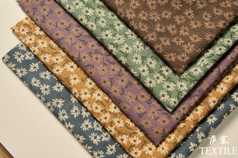 Peut inclure: Une s&eacute;lection de tissus &agrave; motifs floraux dans diverses couleurs, notamment le bleu, l'or, le violet, le vert et le marron. Chaque tissu pr&eacute;sente un motif de marguerites r&eacute;p&eacute;t&eacute;es en blanc ou cr&egrave;me. Les tissus sont dispos&eacute;s en diagonale.