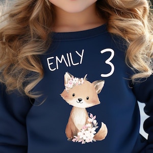 Könnte beinhalten: Ein junges Mädchen trägt ein marineblaues Sweatshirt mit einem Fuchsgrafik, dem Namen "EMILY" und der Zahl "3". Sie trägt auch eine rosa Strickmütze mit Bommel. Der Hintergrund zeigt eine schneebedeckte Berglandschaft.