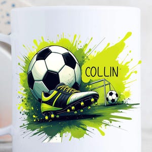 May include: White ceramic mug featuring a soccer theme. The design includes a soccer ball, a soccer cleat, a small goal, and the name "COLLIN" in green. The background has green and yellow paint splatters.