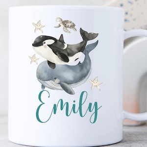 Könnte beinhalten: Weiße Keramik-Tasse mit einer Aquarell-Illustration eines Wals, Orcas, einer Meeresschildkröte und Seesternen. Der Name "Emily" ist in türkisfarbener Schrift geschrieben. Die Tasse hat einen weißen Henkel.