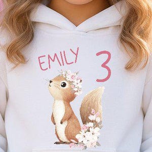 Könnte beinhalten: Ein weißer Kapuzenpullover mit einem Eichhörnchen-Motiv, dem Namen "Emily" und der Zahl "3" in Rosa. Das Mädchen auf dem Bild trägt eine weiße Mütze und eine rosa Hose.