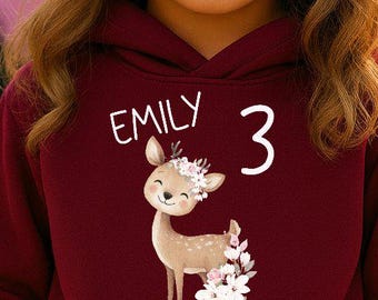 Pode incluir: Uma criança usando um moletom vinho e um gorro combinando. O moletom apresenta um desenho de veado com o nome "EMILY" e o número "3". A criança também está usando calças brancas.