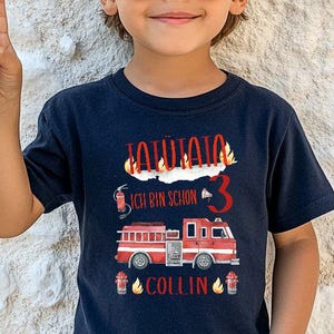 Könnte beinhalten: Marineblaues T-Shirt mit einem roten Feuerwehrauto-Motiv, Flammen und dem Text "Ich bin schon 3" und "Collin". Das T-Shirt ist für den Geburtstag eines Kindes. Das Kind trägt eine Sonnenbrille und zeigt das Friedenszeichen.