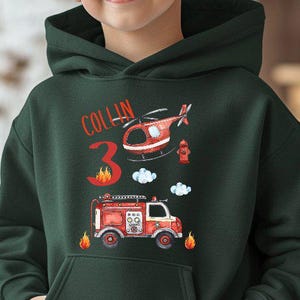 Puede incluir: Una sudadera con capucha verde oscuro con un gráfico colorido de un camión de bomberos, un helicóptero y el número 3. El nombre "Collin" está impreso encima del número. La sudadera tiene un bolsillo delantero.