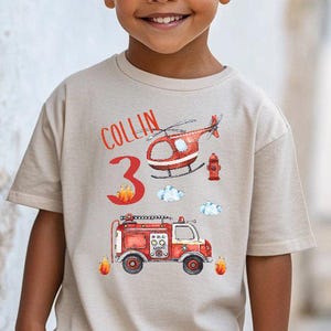 Può includere: Una maglietta beige con un disegno di camion dei pompieri, elicottero e idrante rosso e bianco. Il nome "Collin" e il numero "3" sono stampati in rosso. La maglietta è indossata da un bambino.