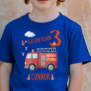 May include: A blue t-shirt featuring a red fire engine design, the number 3, and the name "Connor." The shirt also has the text "Ich bin schon." The boy is wearing blue jeans.