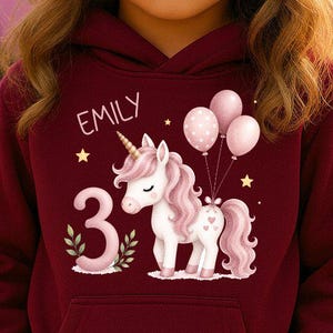 Könnte beinhalten: Ein kastanienbrauner Kapuzenpullover mit einem Grafikdruck eines weißen Einhorns mit rosa Mähne, der Zahl 3, drei rosa Luftballons und dem Namen EMILY in weißer Schrift. Der Hoodie hat eine Kängurutasche und lange Ärmel.