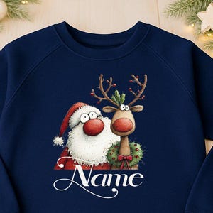 Może przedstawiać: Granatowa bluza z zabawną grafiką Świętego Mikołaja i renifera. Ilustracja zawiera czapkę Świętego Mikołaja, rogi renifera i wieniec. Słowo "Name" jest napisane kursywą. Przytulny element garderoby na sezon świąteczny.