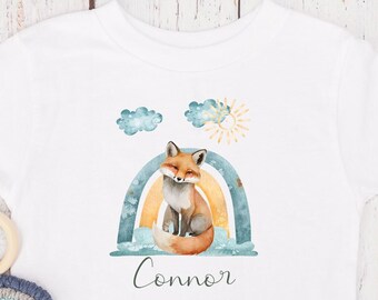 Könnte beinhalten: Weißes Baby-T-Shirt mit einem Fuchs, der auf einem Regenbogen sitzt. Der Regenbogen ist blau, gelb und orange. Über dem Fuchs befinden sich Wolken und eine Sonne. Der Name "Connor" ist unter dem Regenbogen gedruckt.
