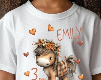 Könnte beinhalten: Weißes T-Shirt mit einem Cartoon-Pony mit Blumenkrone und karierter Decke. Auf dem Shirt stehen der Name "EMILY" und die Zahl "3", mit Herz-Akzenten. Das Shirt ist für Kinder.