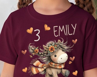 Könnte beinhalten: Ein weinrotes T-Shirt mit einem Cartoon-Pferd, einer Blumenmähne und einem Schal. Die Zahl "3" und der Name "EMILY" sind ebenfalls aufgedruckt. Ein junges Mädchen mit Zöpfen trägt das T-Shirt.