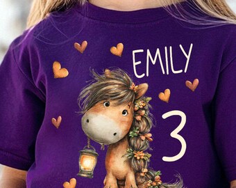 Könnte beinhalten: Ein lila T-Shirt mit einem Cartoon-Pferd, das eine Laterne hält, mit dem Namen "EMILY" und der Zahl "3". Das Shirt hat Herzgrafiken. Das Mädchen trägt eine Sonnenbrille.