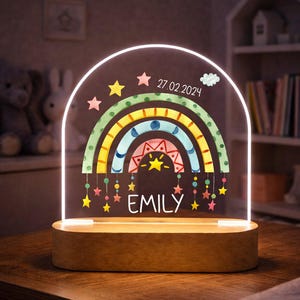 May include: A rainbow-shaped night light with the name "EMILY" and the date "27.02.2024" displayed. The light has a wooden base and a clear acrylic front with colorful rainbow and star designs. A decorative item for a child's room.
