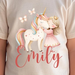 Pu&ograve; includere: Maglietta beige con un'illustrazione di un unicorno e una bambina che si abbracciano. L'unicorno ha una criniera e una coda arcobaleno, e la bambina indossa un vestito rosa. Il nome "Emily" &egrave; scritto in corsivo sotto l'illustrazione.