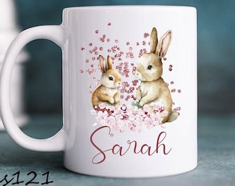 May include: White ceramic mug with a pink floral design and two brown bunnies. The mug has the name "Sarah" written in pink cursive.