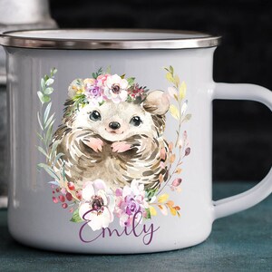 K&ouml;nnte beinhalten: Graue Email Tasse mit einer Aquarell-Illustration eines Igels, der eine Blumenkrone tr&auml;gt. Der Igel ist von Blumen und Gr&uuml;n umgeben. Die Tasse hat einen silbernen Rand und einen Henkel. Der Name "Emily" ist in Lila unter der Illustration gedruckt.
