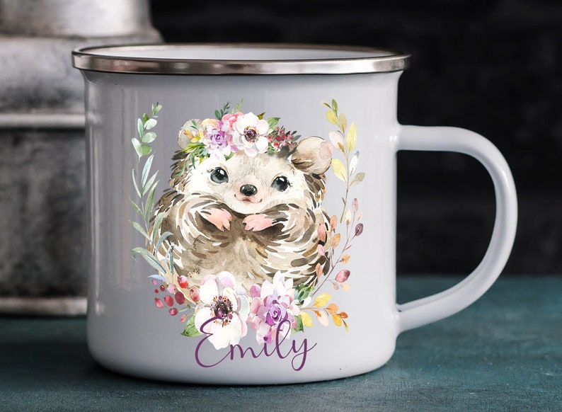 K&ouml;nnte beinhalten: Graue Email Tasse mit einer Aquarell-Illustration eines Igels, der eine Blumenkrone tr&auml;gt. Der Igel ist von Blumen und Gr&uuml;n umgeben. Die Tasse hat einen silbernen Rand und einen Henkel. Der Name "Emily" ist in Lila unter der Illustration gedruckt.