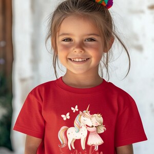Pu&ograve; includere: Una bambina indossa una maglietta rossa con una grafica colorata di un unicorno, una principessa e farfalle. Il nome "Emily" &egrave; scritto sotto. Ha un accessorio per capelli colorato.