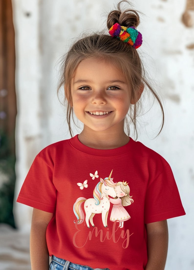 Pu&ograve; includere: Una bambina indossa una maglietta rossa con una grafica colorata di un unicorno, una principessa e farfalle. Il nome "Emily" &egrave; scritto sotto. Ha un accessorio per capelli colorato.