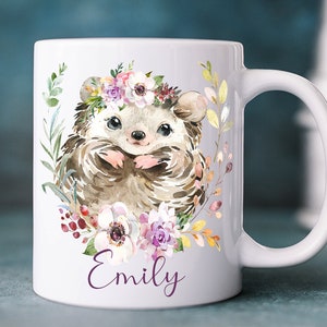 K&ouml;nnte beinhalten: Wei&szlig;e Keramiktasse mit einer Aquarell-Illustration eines Igels, der eine Blumenkrone tr&auml;gt. Der Igel ist von Blumen und Bl&auml;ttern umgeben. Die Tasse tr&auml;gt den Namen "Emily" auf dem Boden.