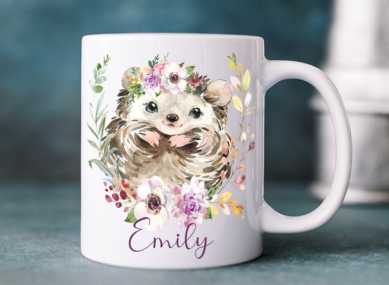 K&ouml;nnte beinhalten: Wei&szlig;e Keramiktasse mit einer Aquarell-Illustration eines Igels, der eine Blumenkrone tr&auml;gt. Der Igel ist von Blumen und Bl&auml;ttern umgeben. Die Tasse tr&auml;gt den Namen "Emily" auf dem Boden.