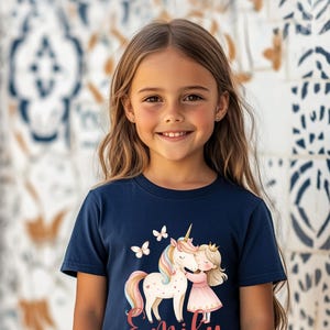 Pu&ograve; includere: T-shirt blu navy con un design di unicorno, una principessa e farfalle. Il nome "Emily" &egrave; scritto in corsivo. La maglietta ha un girocollo e maniche corte.