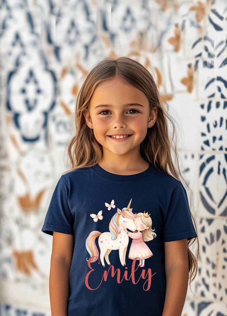 Pu&ograve; includere: T-shirt blu navy con un design di unicorno, una principessa e farfalle. Il nome "Emily" &egrave; scritto in corsivo. La maglietta ha un girocollo e maniche corte.