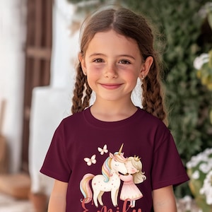 Pu&ograve; includere: Maglietta bordeaux con un design stravagante di un unicorno, una ragazza e farfalle. L'unicorno ha una criniera e una coda arcobaleno. La ragazza indossa un vestito rosa e una corona. Il nome "Emily" &egrave; scritto in corsivo.