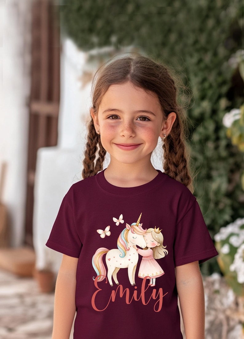 Pu&ograve; includere: Maglietta bordeaux con un design stravagante di un unicorno, una ragazza e farfalle. L'unicorno ha una criniera e una coda arcobaleno. La ragazza indossa un vestito rosa e una corona. Il nome "Emily" &egrave; scritto in corsivo.