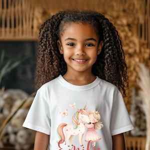 Pu&ograve; includere: T-shirt bianca con un'illustrazione colorata di un unicorno, una ragazza e farfalle. L'unicorno ha un corno d'oro e una criniera e coda arcobaleno. La ragazza indossa un vestito rosa e una corona. Il nome "Emily" &egrave; scritto in corsivo.