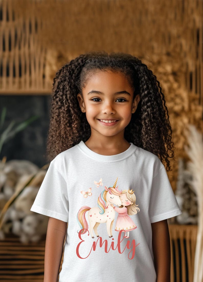 Pu&ograve; includere: T-shirt bianca con un'illustrazione colorata di un unicorno, una ragazza e farfalle. L'unicorno ha un corno d'oro e una criniera e coda arcobaleno. La ragazza indossa un vestito rosa e una corona. Il nome "Emily" &egrave; scritto in corsivo.