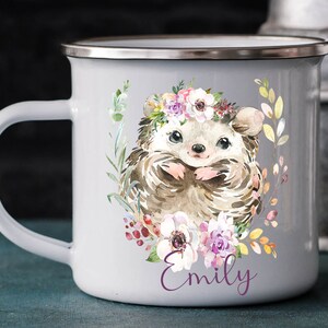 K&ouml;nnte beinhalten: Eine wei&szlig;e Emailletasse mit einer Aquarell-Illustration eines Igels, der eine Blumenkrone tr&auml;gt. Der Igel ist von Blumen und Bl&auml;ttern umgeben. Die Tasse hat einen silbernen Rand und einen Henkel. Der Name "Emily" ist in Lila auf die Tasse gedruckt.