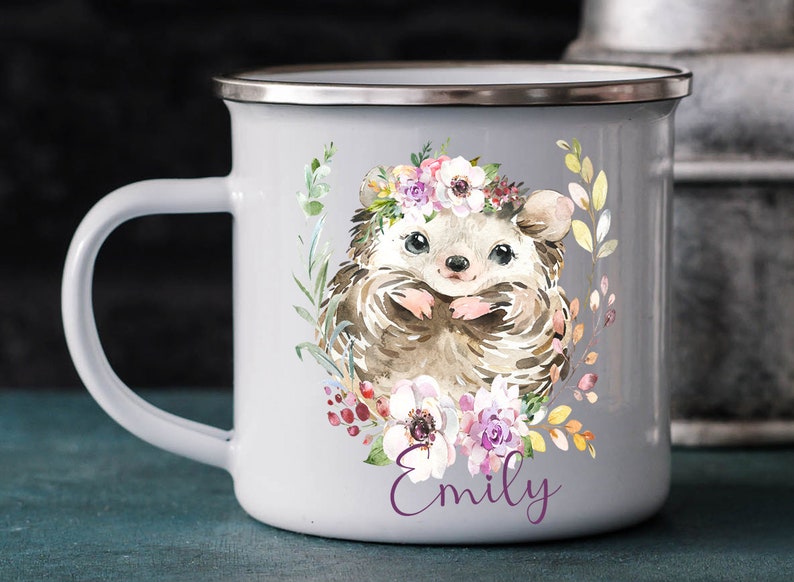 K&ouml;nnte beinhalten: Eine wei&szlig;e Emailletasse mit einer Aquarell-Illustration eines Igels, der eine Blumenkrone tr&auml;gt. Der Igel ist von Blumen und Bl&auml;ttern umgeben. Die Tasse hat einen silbernen Rand und einen Henkel. Der Name "Emily" ist in Lila auf die Tasse gedruckt.