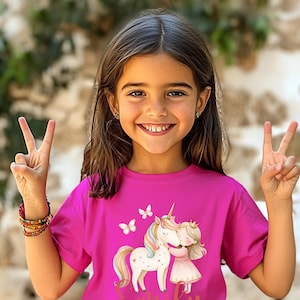 Pu&ograve; includere: Una maglietta magenta con un disegno di unicorno e bambina che si abbracciano, farfalle e il nome "Emily" in oro. La ragazza sorride e fa il segno della vittoria.