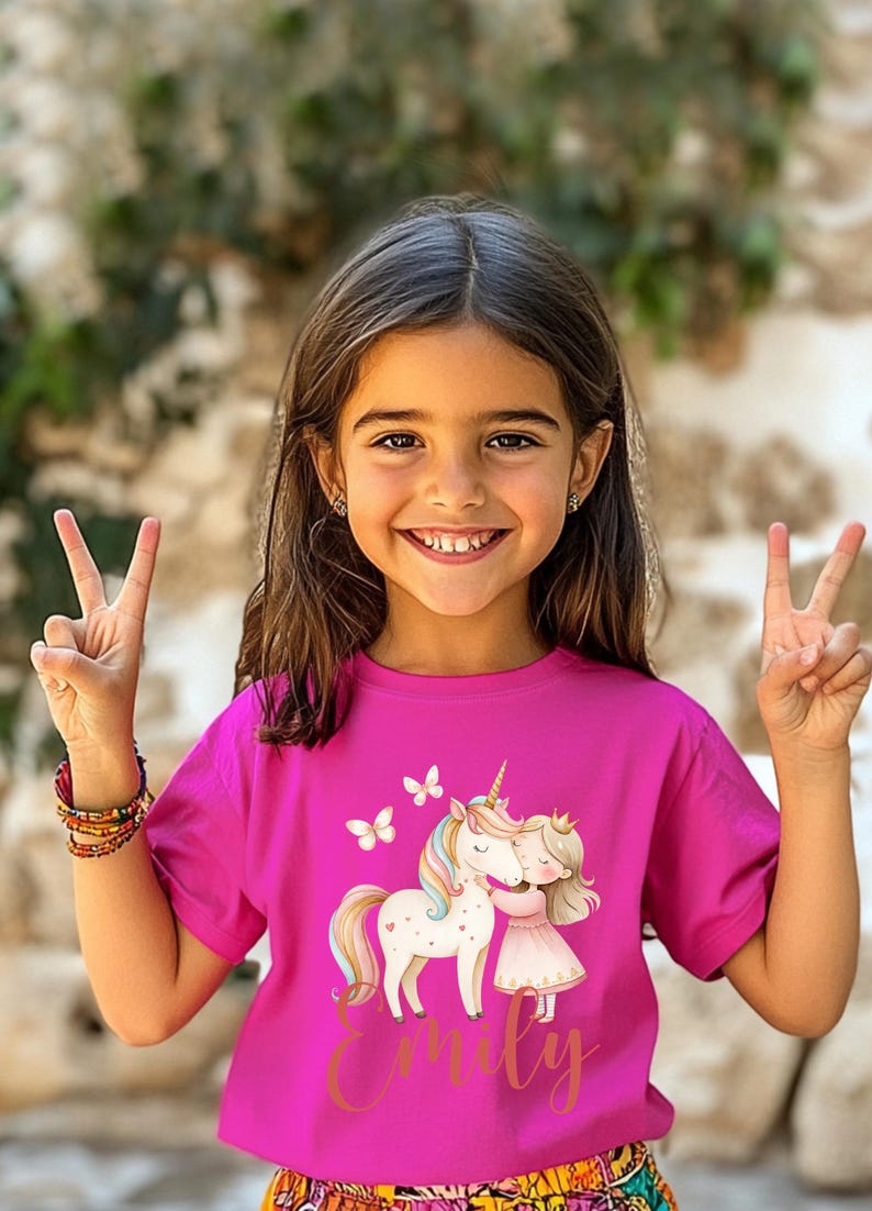 Pu&ograve; includere: Una maglietta magenta con un disegno di unicorno e bambina che si abbracciano, farfalle e il nome "Emily" in oro. La ragazza sorride e fa il segno della vittoria.
