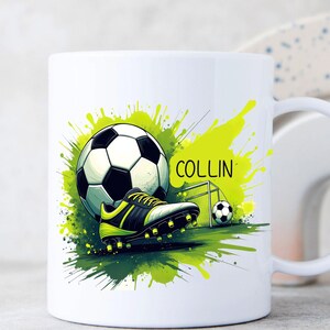 May include: White ceramic mug featuring a soccer ball, cleats, and a small goal, with the name "COLLIN" in green. The design includes green and yellow paint splatters, creating a dynamic sports-themed look.