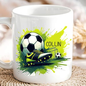May include: White ceramic mug featuring a soccer theme. The design includes a soccer ball, cleats, and a small goal, set against a green splash background. The name "COLLIN" is printed on the mug.
