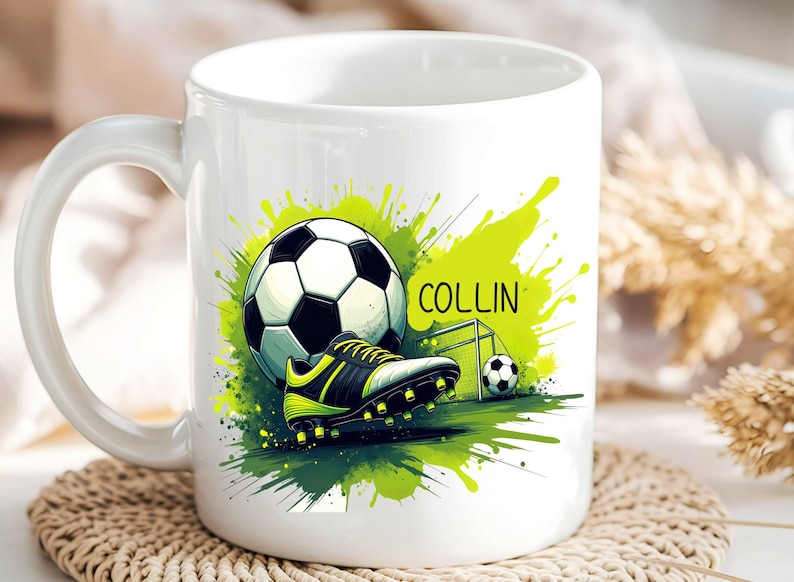 May include: White ceramic mug featuring a soccer theme. The design includes a soccer ball, cleats, and a small goal, set against a green splash background. The name "COLLIN" is printed on the mug.