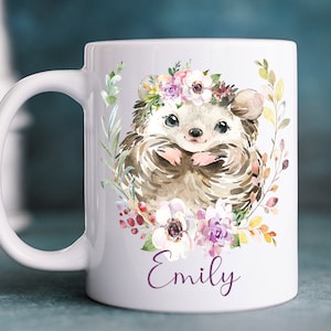 K&ouml;nnte beinhalten: Wei&szlig;e Keramiktasse mit einer Aquarell-Illustration eines Igels, der eine Blumenkrone tr&auml;gt. Der Igel ist von Blumen und Bl&auml;ttern umgeben. Die Tasse tr&auml;gt den Namen "Emily" in Lila unten aufgedruckt.