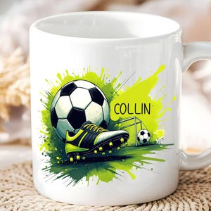 May include: White ceramic mug featuring a soccer theme. The design includes a soccer ball, cleats, and a goal, with a green and yellow paint splash effect. The name "COLLIN" is printed on the mug.