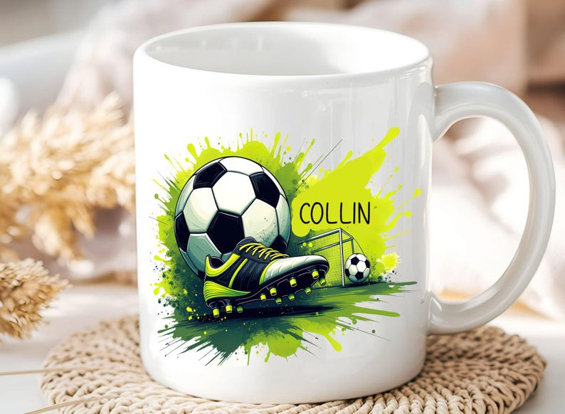 May include: White ceramic mug featuring a soccer theme. The design includes a soccer ball, cleats, and a goal, with a green and yellow paint splash effect. The name "COLLIN" is printed on the mug.