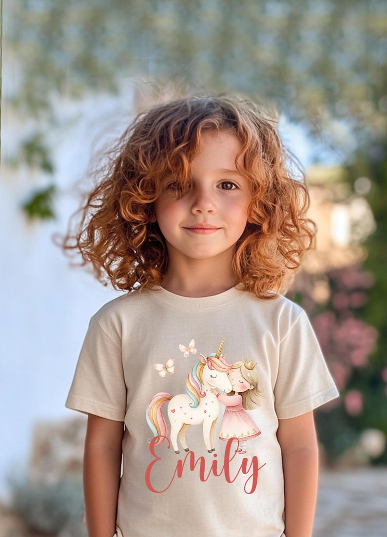 Pu&ograve; includere: Maglietta beige con un'illustrazione di un unicorno e una bambina che si abbracciano. L'unicorno ha una criniera e una coda arcobaleno, e la bambina indossa un vestito rosa. Il nome "Emily" &egrave; scritto in corsivo sotto l'illustrazione.