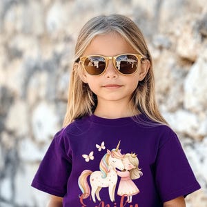 Pu&ograve; includere: Una maglietta viola per bambini con un disegno di unicorno e principessa, con il nome "Emily" in corsivo. La persona indossa occhiali da sole con montatura dorata. Sfondo strutturato.